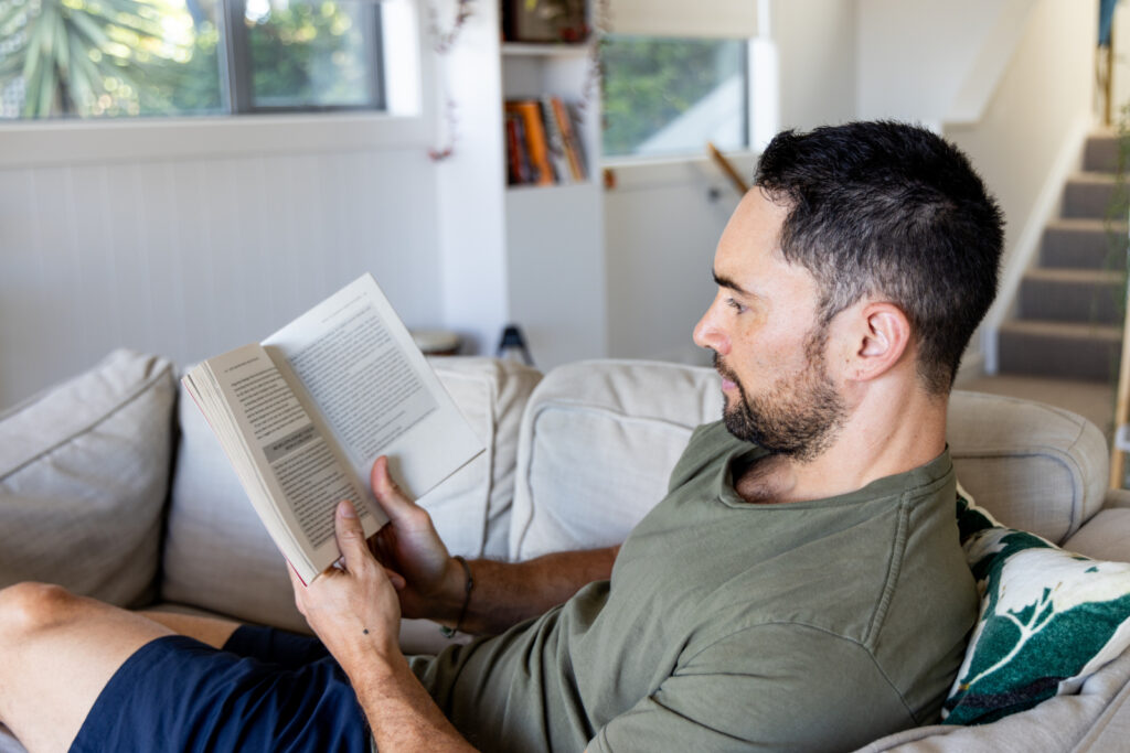 Man reading a book while resting on a couch, stairs are in the background.